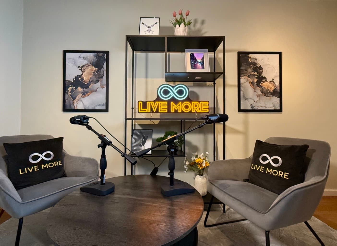 Podcast studio with grey armchairs, branded pillows, microphones, and a glowing 'LIVE MORE' neon sign.