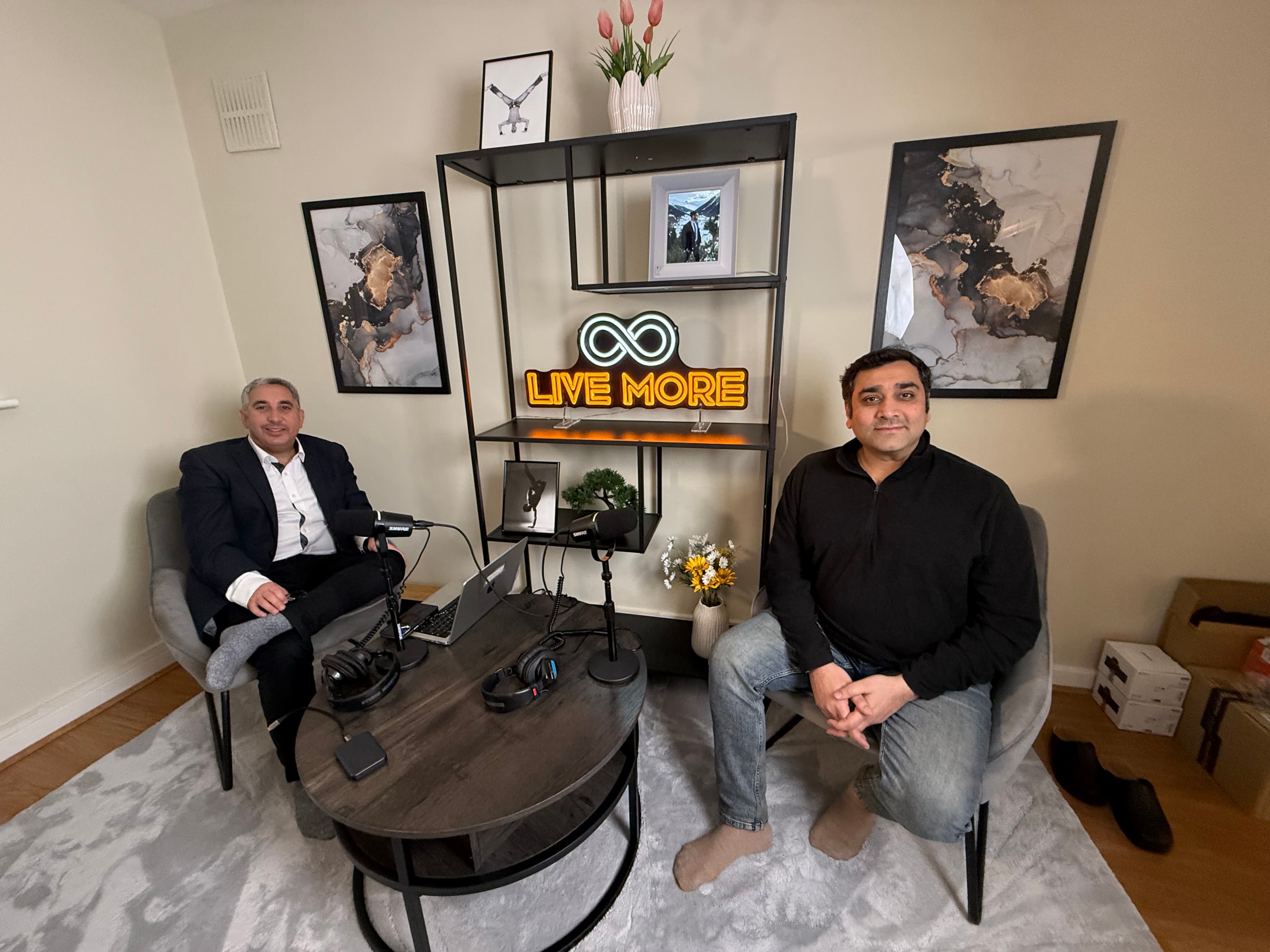 Two men sit in a podcast studio with microphones and a "LIVE MORE" neon sign.