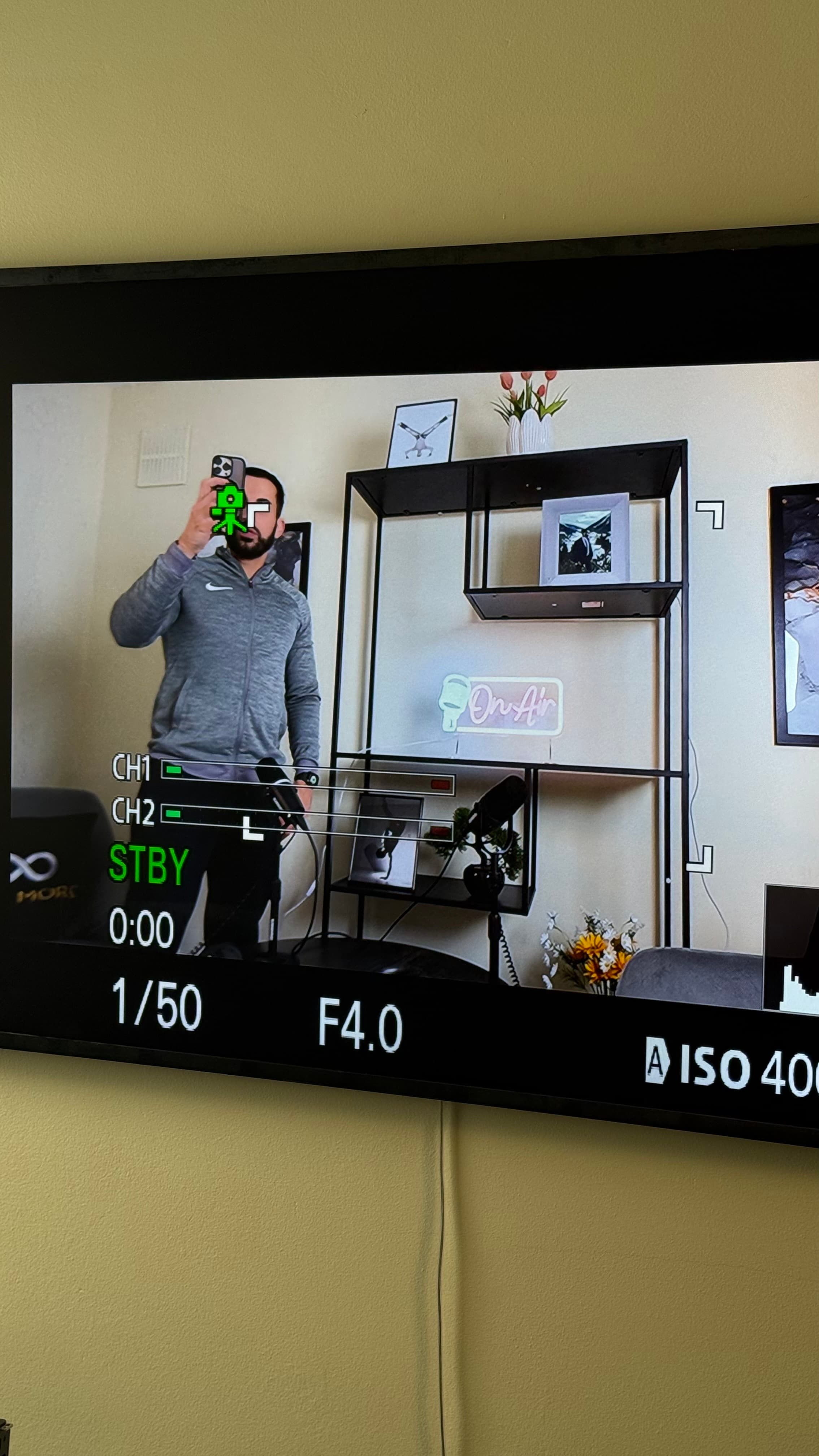Man in grey hoodie filming himself, seen through a camera monitor with technical display overlays.