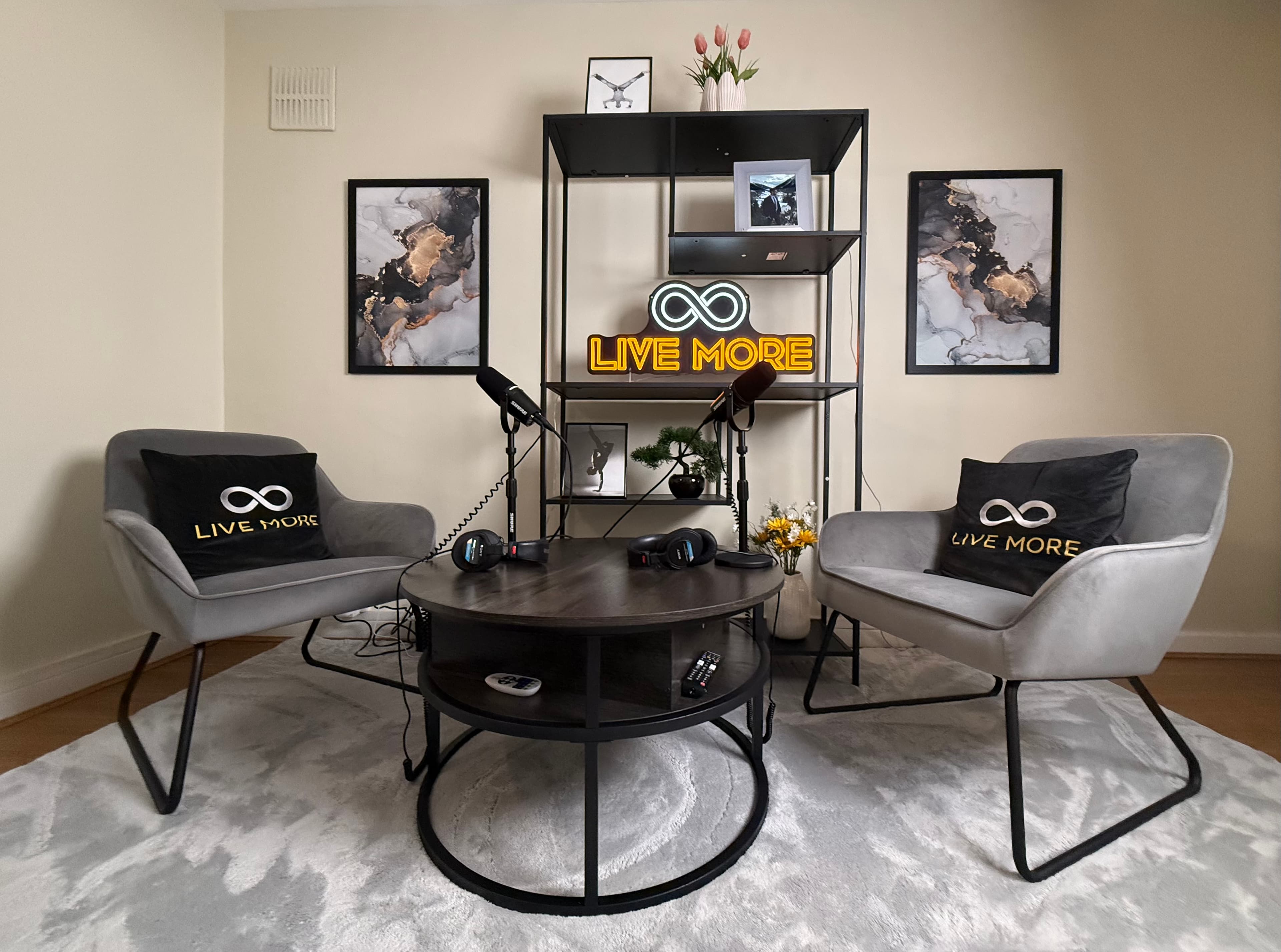 Modern podcast studio featuring grey armchairs, microphones, and a glowing neon 'LIVE MORE' sign.