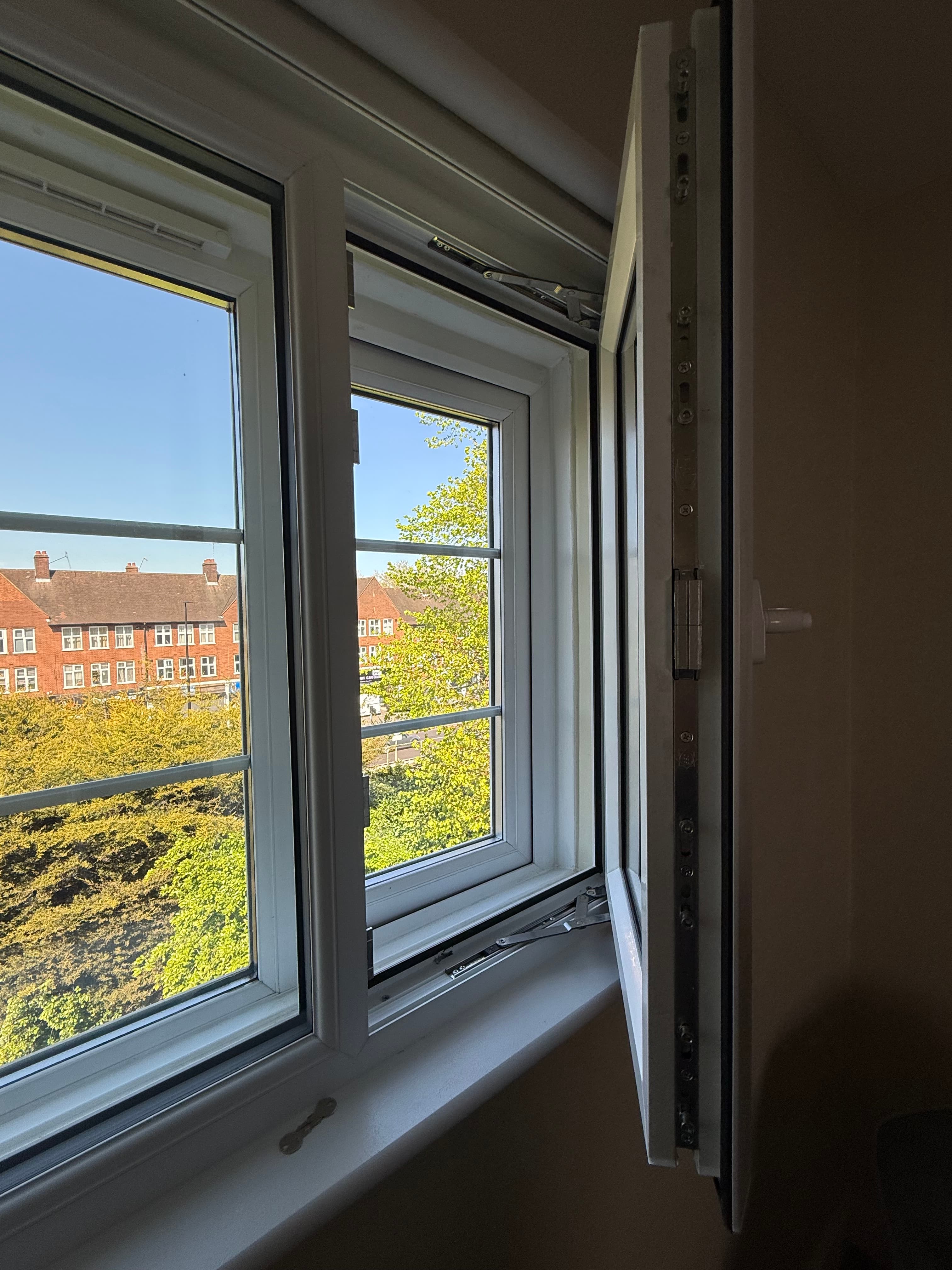 Looking up through an open white window at a brick building and lush green trees.