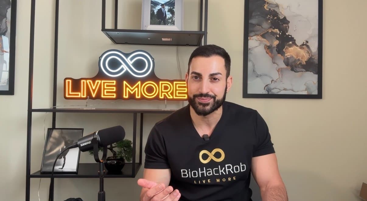 Bearded man in a BioHackRob shirt sits before a neon 'LIVE MORE' sign with microphone.
