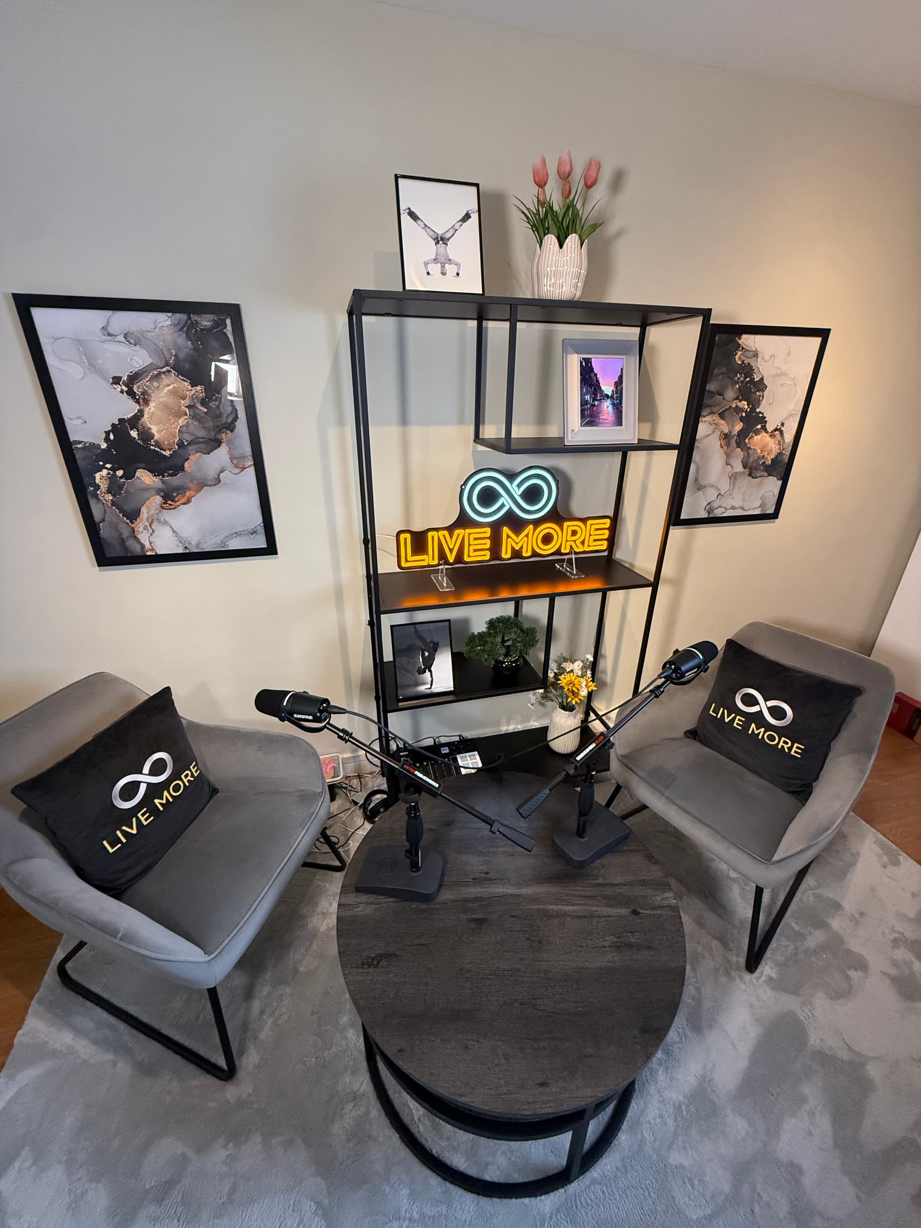 Modern podcast studio with grey armchairs, microphones, a round table, and 'LIVE MORE' neon sign.