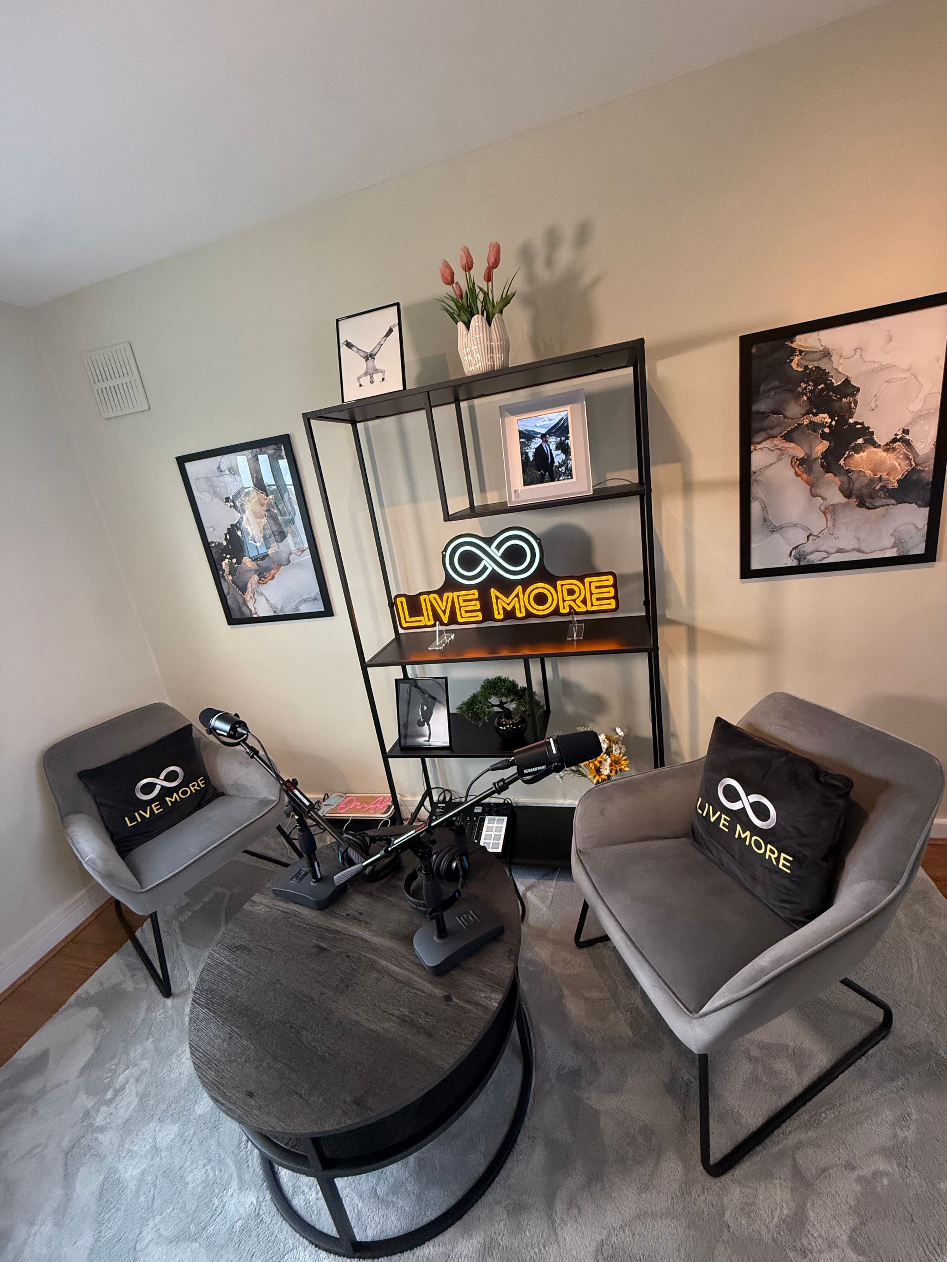 Podcast studio featuring grey armchairs, microphones, and a neon 'LIVE MORE' sign on shelves.