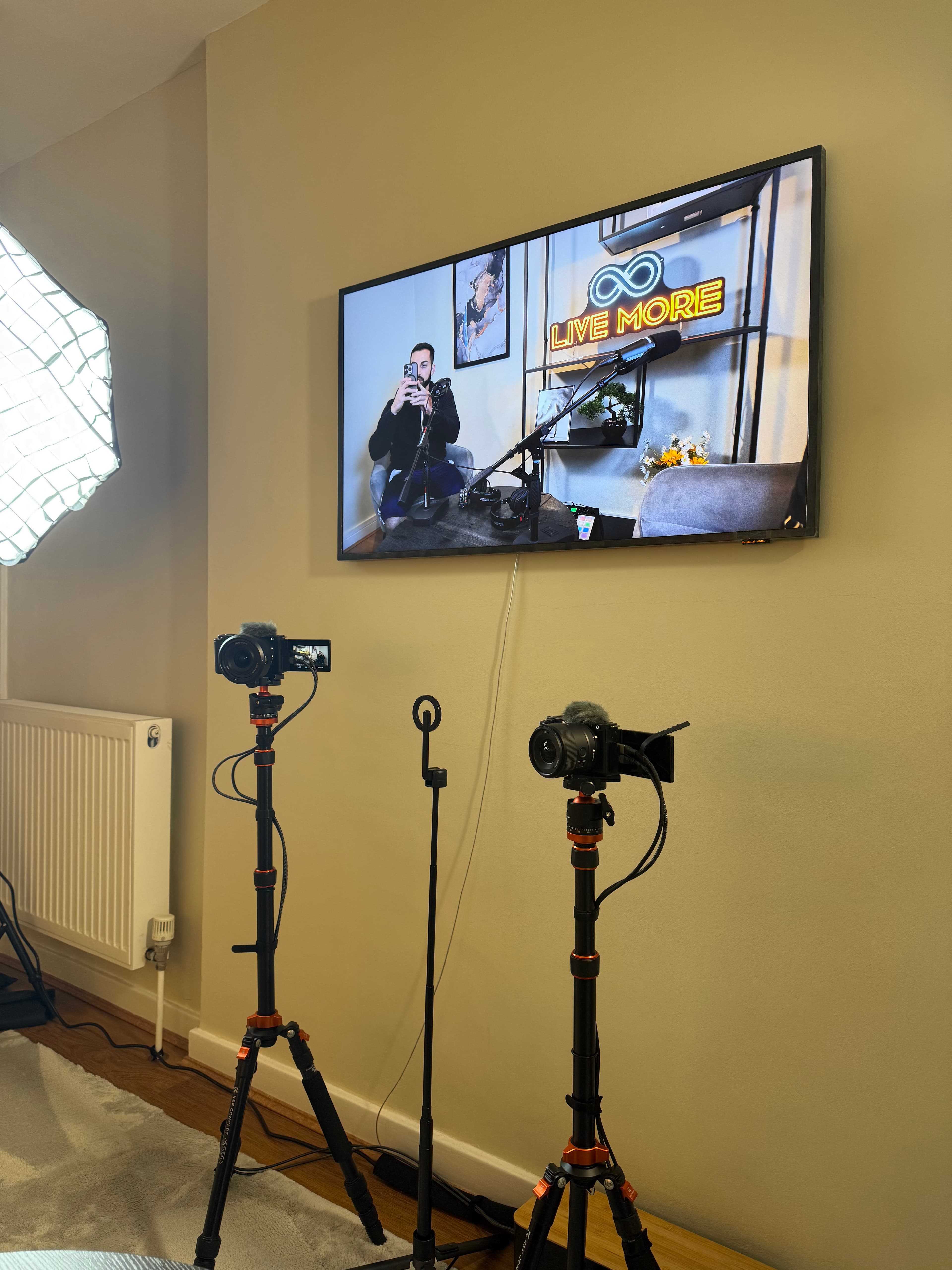 Two cameras on tripods face a wall-mounted monitor showing a live studio recording session.