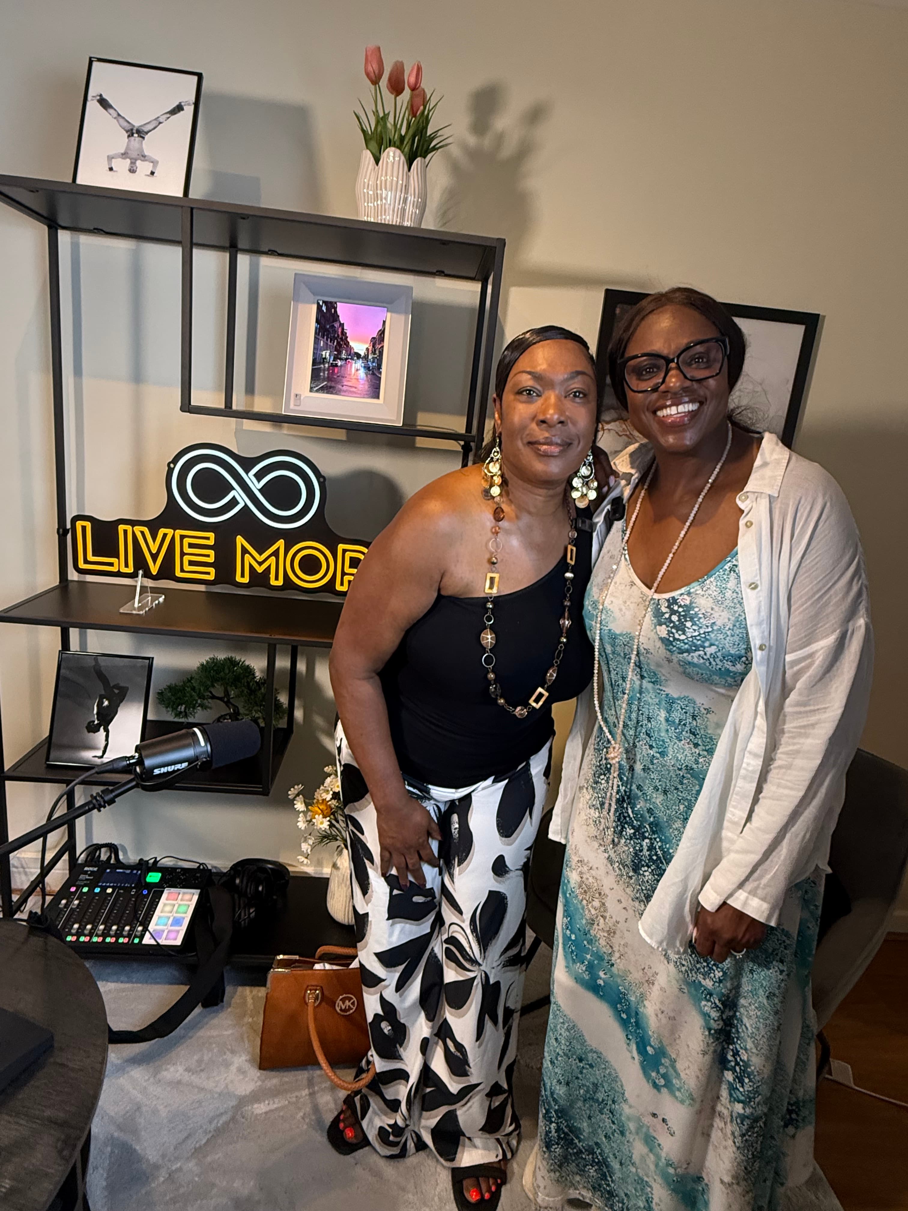 Two smiling Black women pose in a studio with a "LIVE MORE" neon sign.