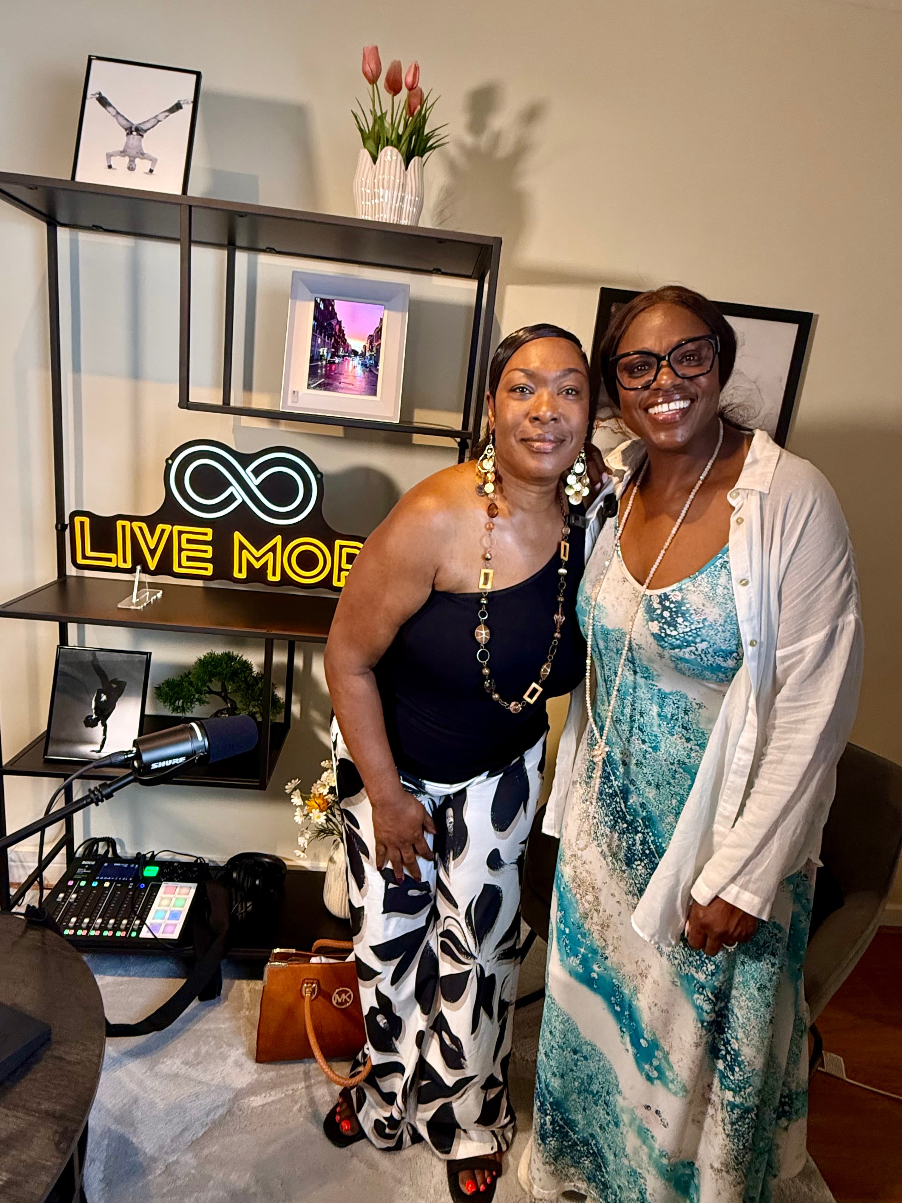 Two smiling Black women pose in a studio with a neon sign and podcast gear.