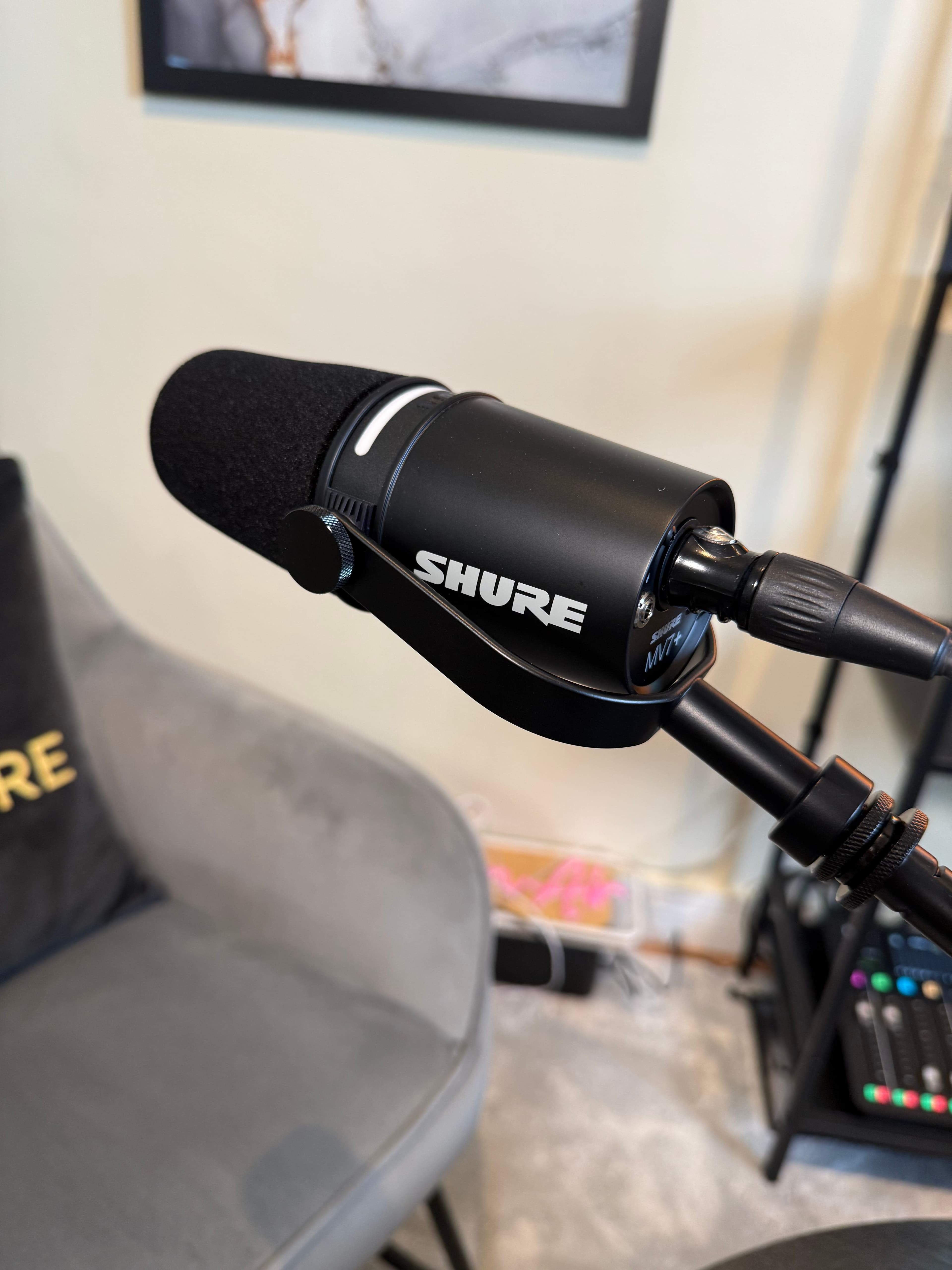 Black Shure MV7+ microphone with foam windscreen mounted on a stand in a home studio.