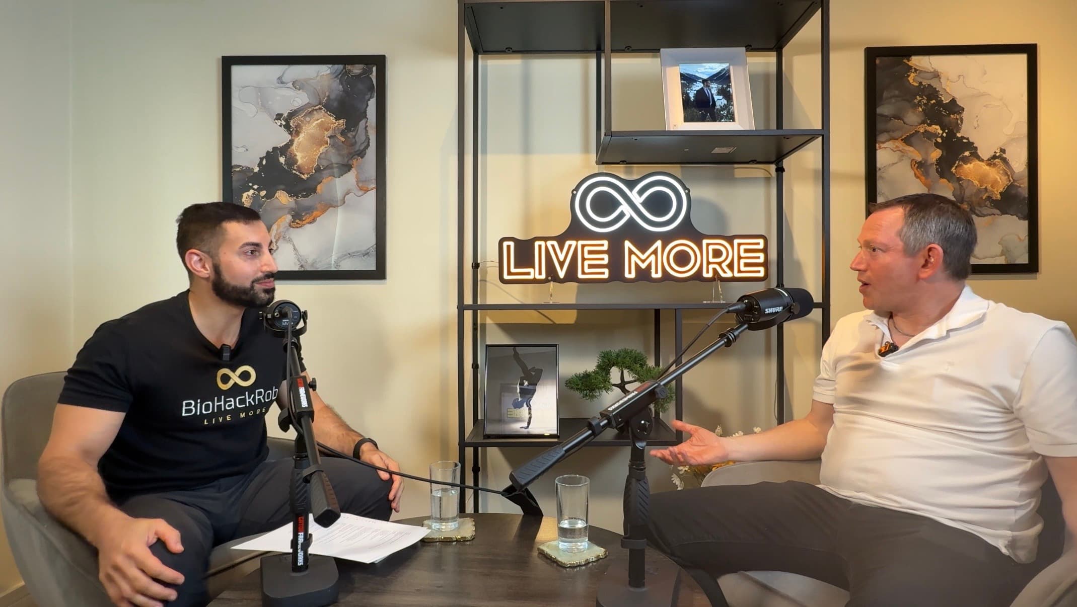 Two men sit in a podcast studio with microphones and a neon 'LIVE MORE' sign.