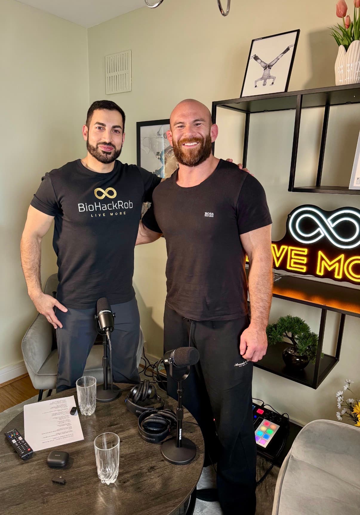 Two muscular men smiling in a podcast studio with microphones and neon 'LIVE MORE' sign.