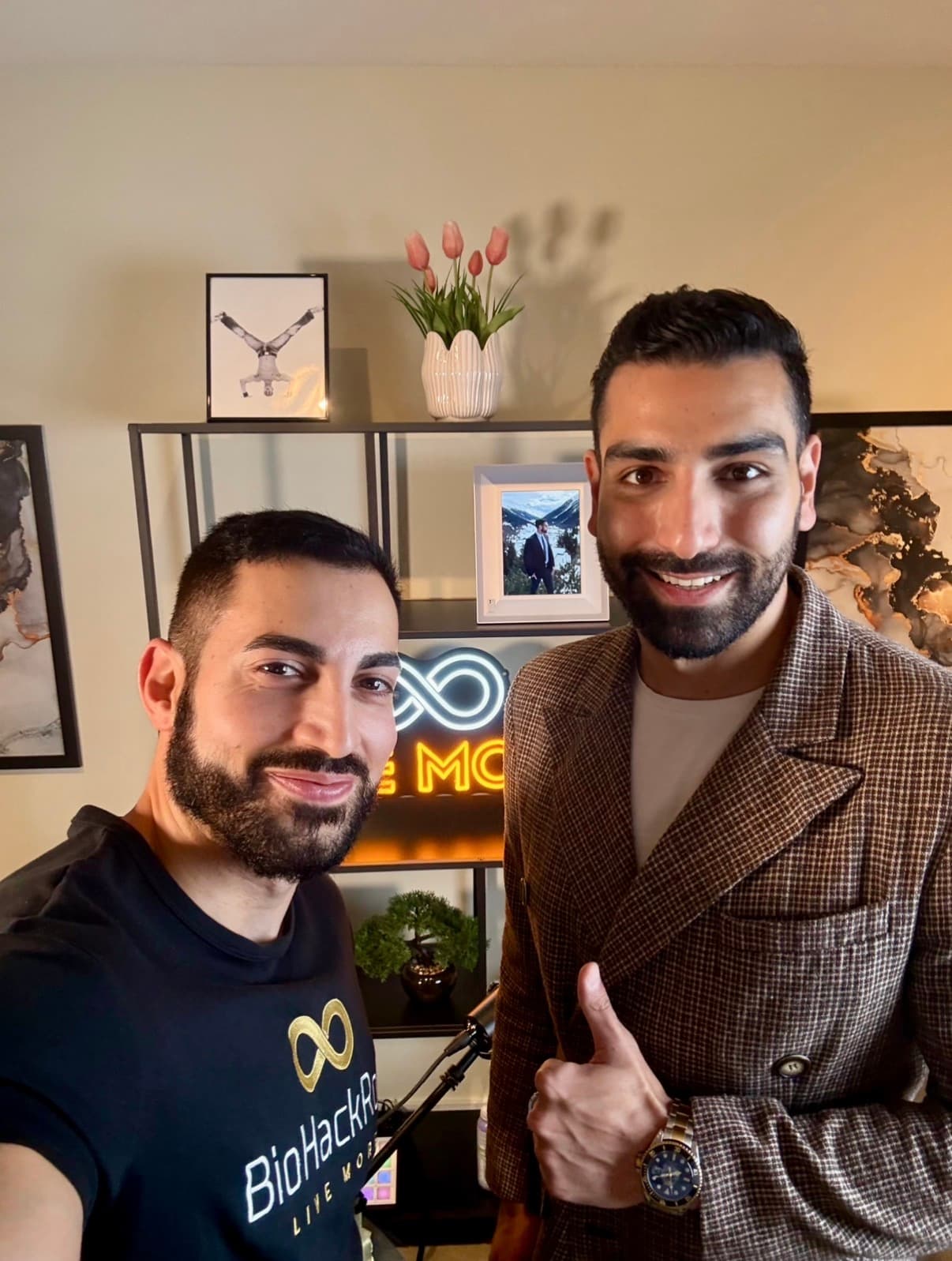 Two bearded men smile in a room, one in a blazer giving a thumbs up.