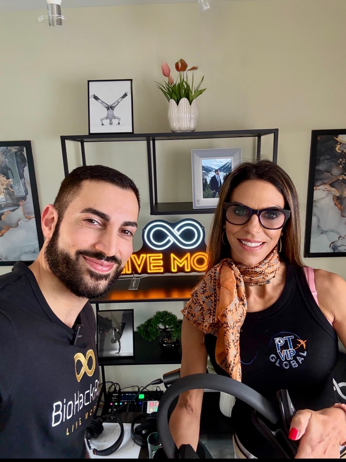 A bearded man and woman with glasses smile before a neon "LIVE MORE" infinity sign.