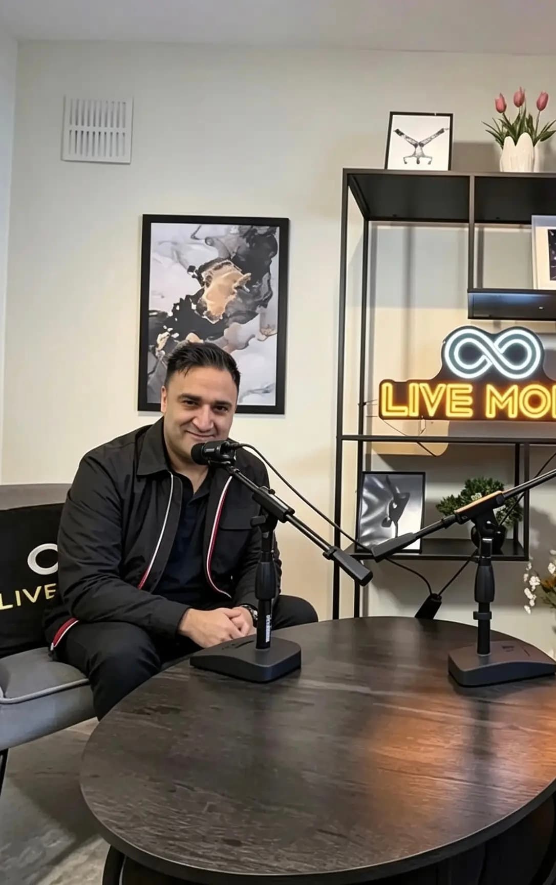 Smiling man at a podcast desk with microphones, featuring a neon 'LIVE MORE' sign.