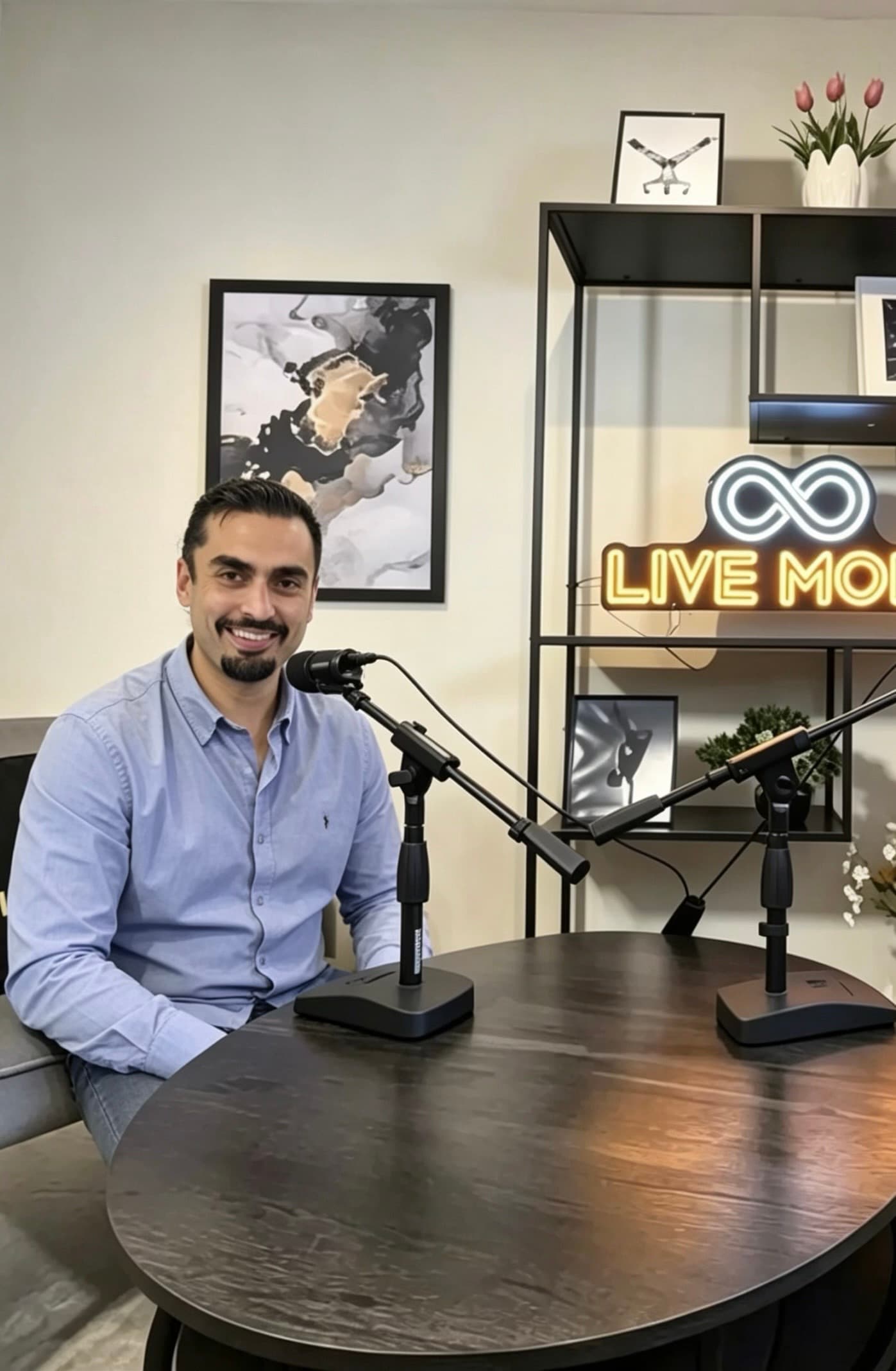 A man with a goatee smiles at a podcast table with two professional microphones.