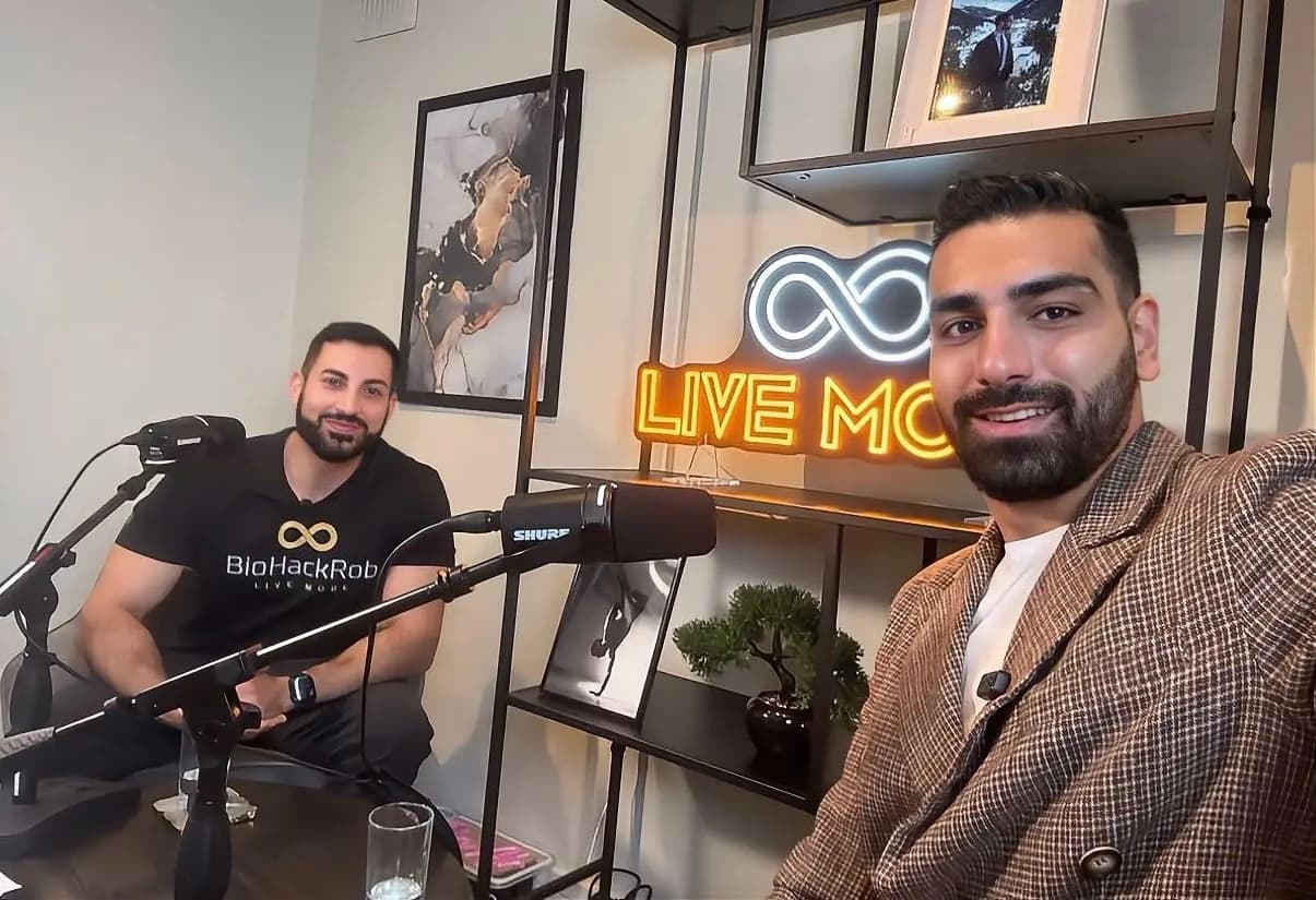 Two men smile in a podcast studio featuring microphones and a neon "LIVE MORE" sign.