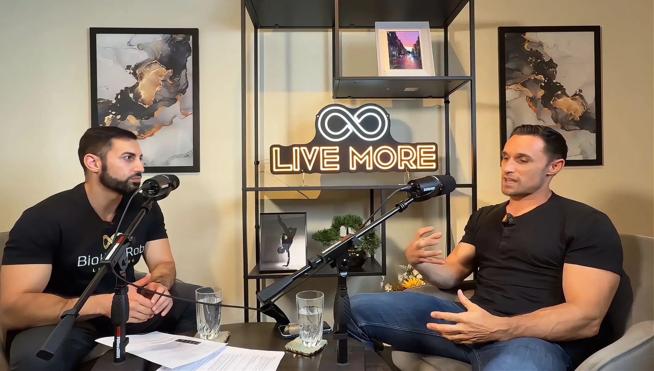 Two men recording a podcast in a studio with a neon "LIVE MORE" sign.